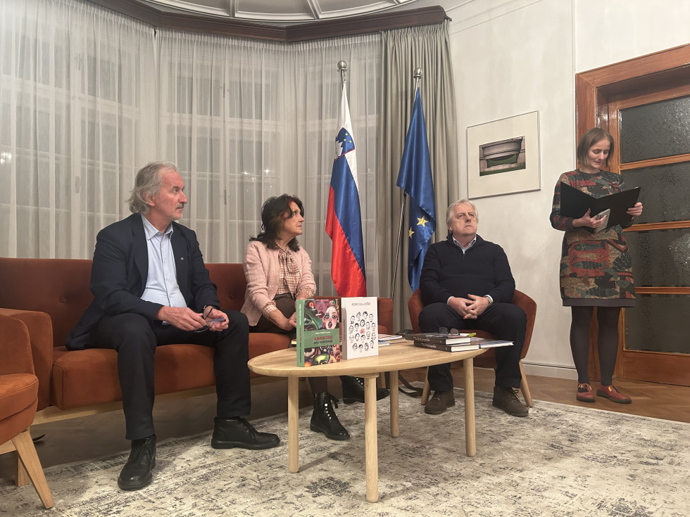 Na fotografiji je notranji prostor. V ospredju je svetla lesena klubska mizica. Na njej sta razstavljeni dve knjigi, obrnjeni tako, da se vidita njuni naslovnici. Okoli mizice sedijo trije ljudje na rjavem kavču in naslanjaču. Povsem desno stoji ženska.