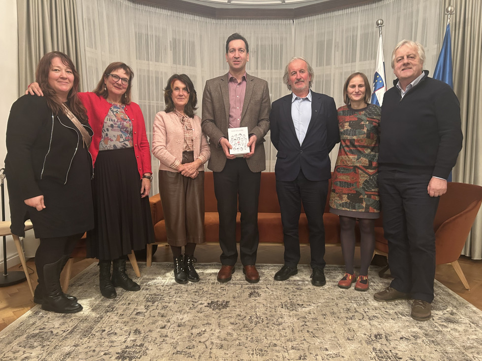 Na fotografiji je skupina sedmih odraslih ljudi, ki stojijo v vrsti in gledajo proti fotografu. Vsi so elegantno oblečeni. V sredini stoji moški v rjavem suknjiču in svetli srajci, ki v rokah drži knjigo s pretežno belimi platnicami. 