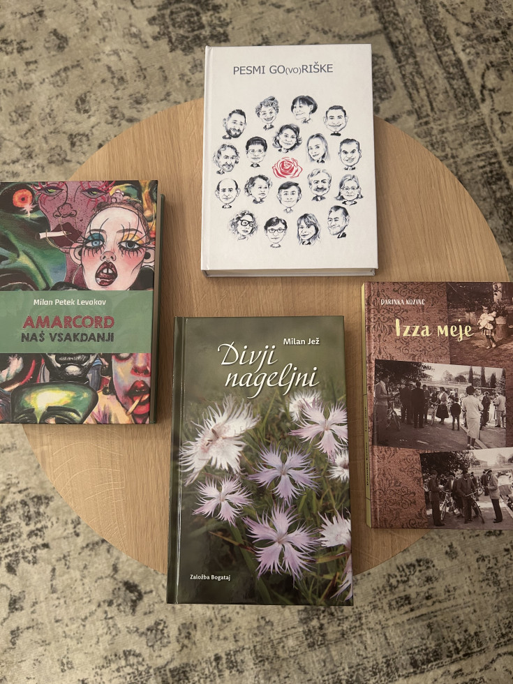 Na fotografiji je pogled od zgoraj na majhno, okroglo leseno mizico. Na mizici so razporejene štiri knjige:Na vrhu, na sredini, leži bela knjiga z naslovom “PESMI GO(VO)RIŠKE”. 