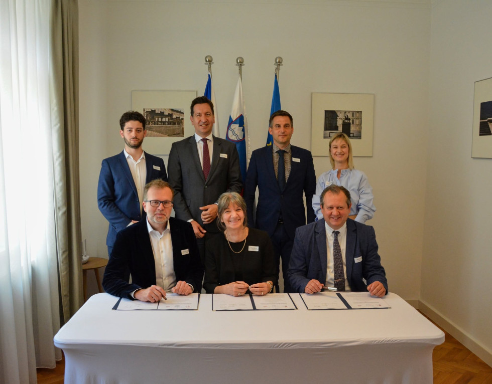 Tri osebe sedijo za mizo, pred njimi pa so odprte mape s podpisanimi dokumenti. Za njimi stojijo še štiri osebe. Vsi so elegantno oblečeni. V ozadju tri zastave, slovenska, češka in zastava Evropske unije, ter dve fotografiji na steni.
