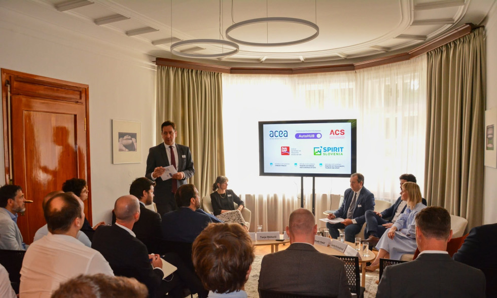 V velikem notranjem prostoru osebe sedijo v polkrogu, moški v temni obleki pa stoji pred njimi in jih nagovarja. Na velikem zaslonu v ozadju so prikazani logotipi organizacij, med njimi ACEA, CEE AutoHUB, ACS in SPIRIT Slovenija. 