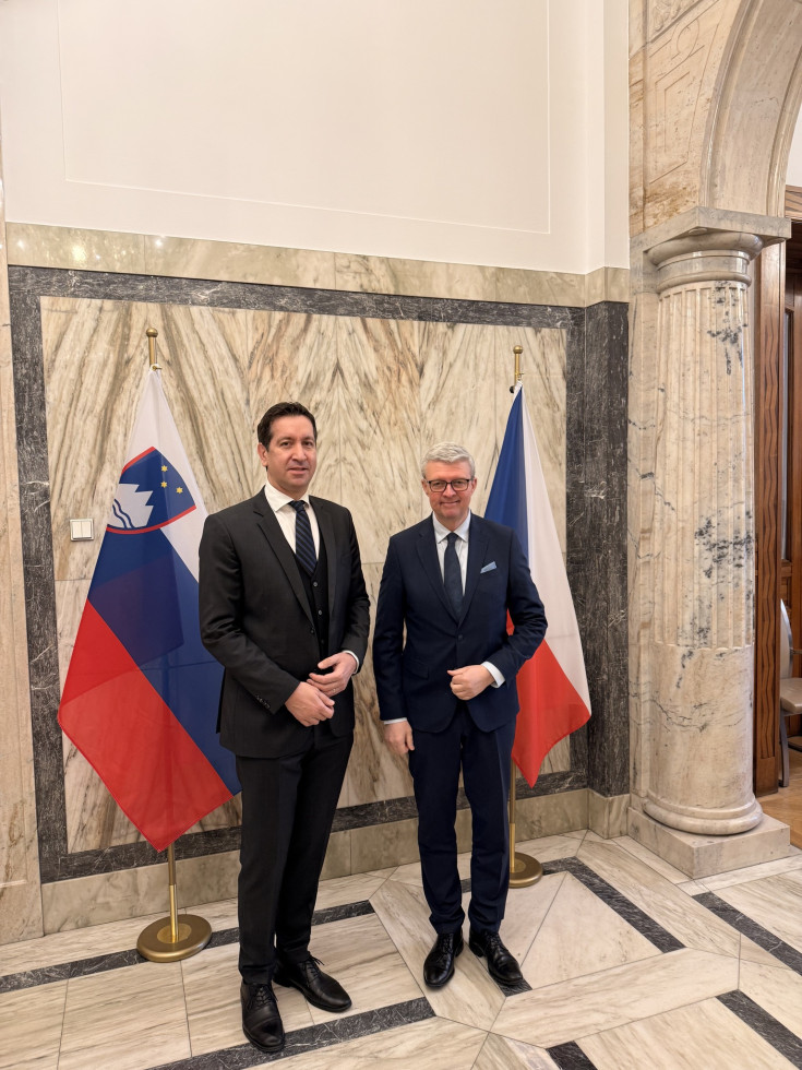 Fotografija dveh moških srednjih let, ki sta oblečena v temni obleki s kravato, ter pozirata pred slovensko in češko zastavo na drogovih.. Prostor v katerem se nahajata je po stenah in tleh obložen s svetlo kamnino. Na desni kamnit steber.