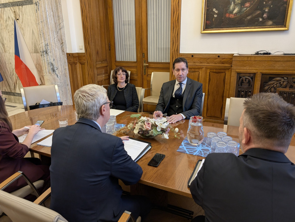 Na fotografiji je skupina petih ljudi, ki sedijo okoli lesene mize in imajo sestanek.