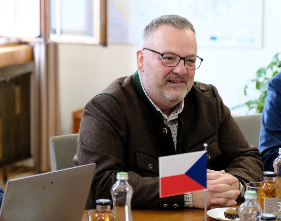 Na fotografiji moški srednjih let z očali, ki sedi za mizo in ima sklenjene roke. Prav tako vidna na fotografiji je češka zastava. 