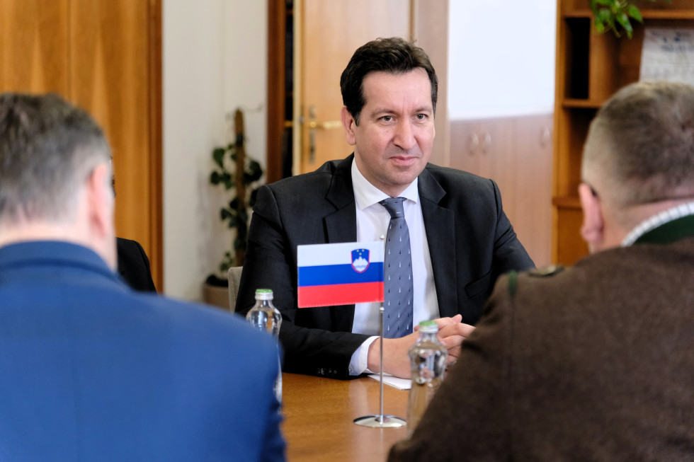 V sredini slike vidimo na mizi majhno slovensko zastavo, za mizo sedi moški srednjih let v črni obleki, beli srajci in modri kravati.