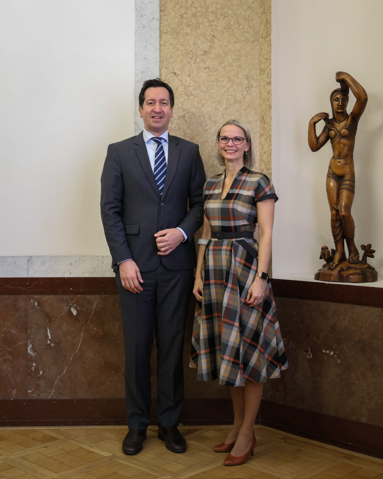 Na fotografiji stojita moški in ženska v notranjem prostoru. Moški stoji na levi. Oblečen je v temno modri suknjič z belo srajco in svetlo modro kravato. Ženska stoji tik ob njem. Oblečena je v karirasto obleko, nosi očala. Oba gledata proti fotografu.