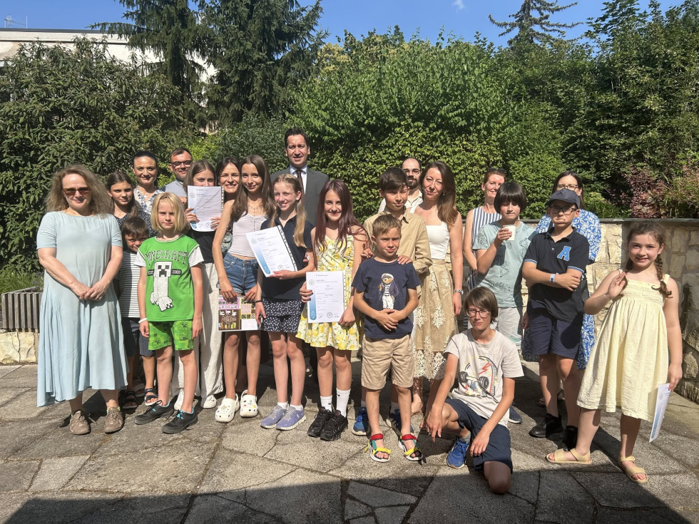 Skupinska fotografija na zunanjem dvorišču. Otroci stojijo in sedijo v ospredju, nekateri v rokah držijo spričevala. Za njimi stojijo odrasli, med njimi veleposlanik v temni obleki in kravati. V ozadju zelenje in modro nebo.
