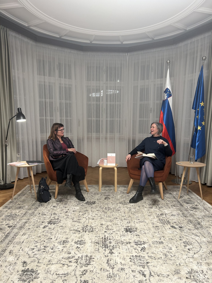 Dve ženski srednjih let v temnih oblačilih sedita na naslanjačih. Poleg imata lesene mizice, na levi strani fotografije je prižgana svetilka, na desni drog z slovensko zastavo in zastavo EU. Na tleh svetel tepih, okna v ozadju zakrita z belimi zavesami.