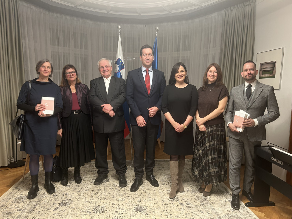 Skupina ljudi pozira za fotografijo. Stojijo v sredini in so obrnjeni proti fotografu. Vsi so elagantno oblečeni. Osebi na skrajni levi in desni strani držita v rokah knjigo roza barve.