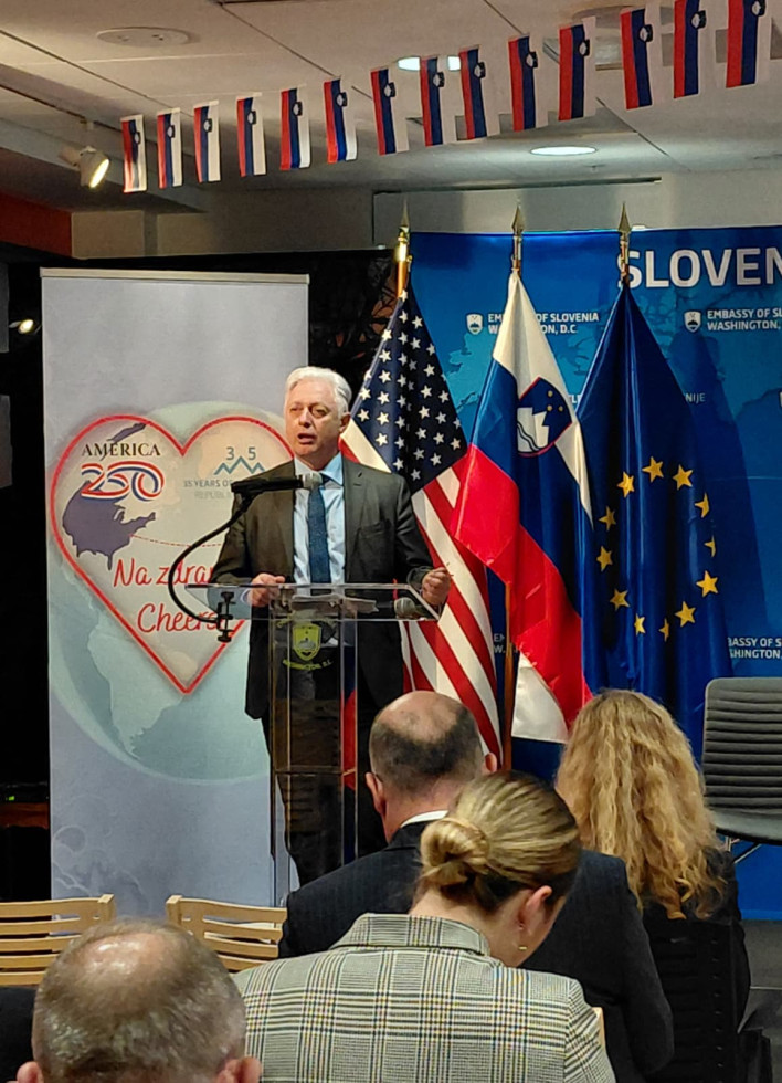 A man speaks to an audience with the American, Slovenian, and EU flags behind him.