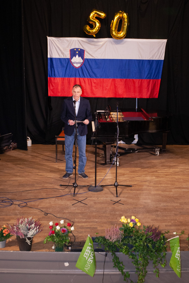 veleposlanik stoji na odru, v ozadju slovenska zastava in baloni v obliki petdesetke