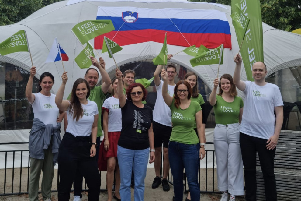 the team is standing and waving flags, in the background a Slovenian pavilion with a large Slovenian flag