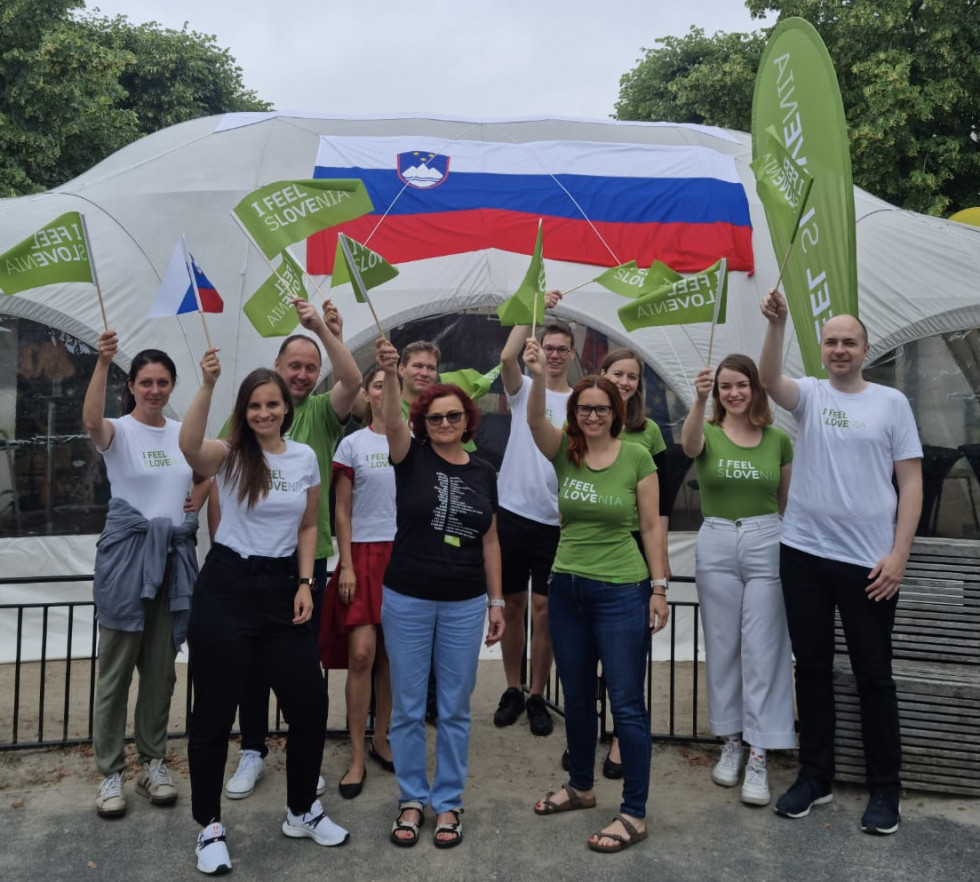 ekipa stoji in maha z zastavicami, v ozadju slovenski paviljon z veliko slovensko zastavo