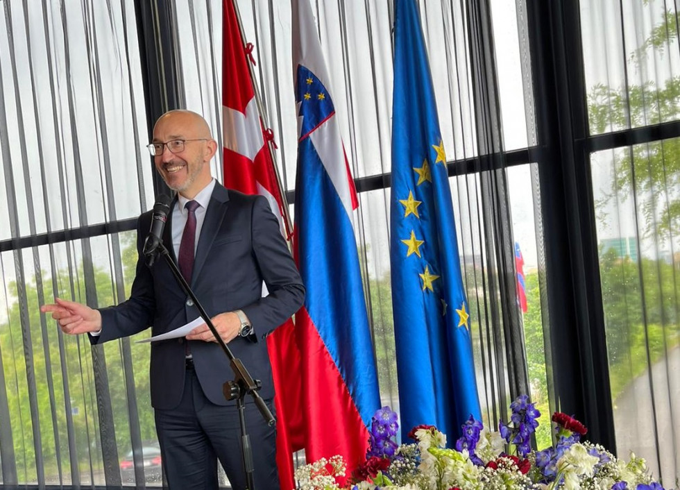 Ambassador stands on the stage, flags in the background, flowers in front