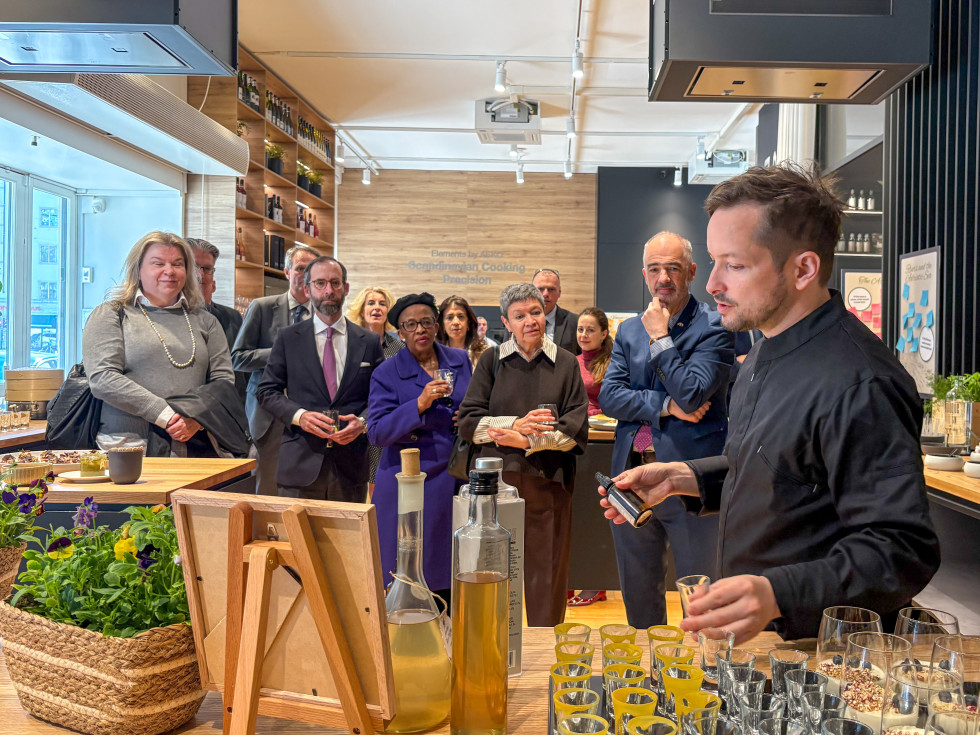 A group of guets stand in a kitchen showroom, attentively listening to a chef who is demonstrating degustation of Slovenian Vodka with hydrolate sprays. 