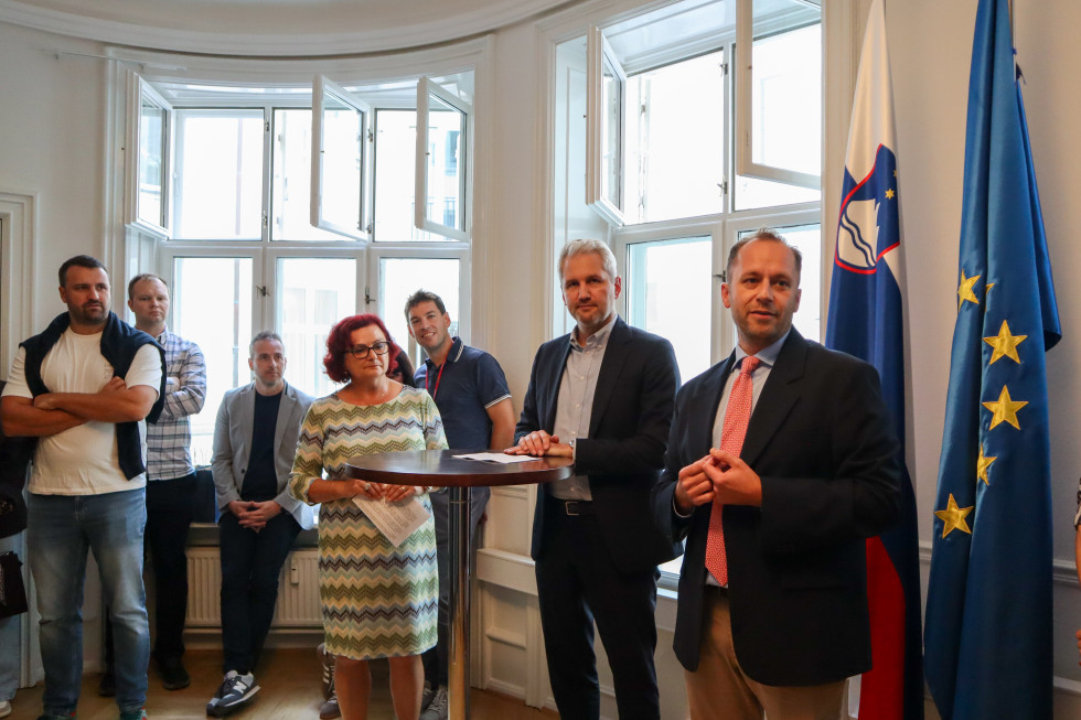 Formally dressed people stand in a room where in the right corner hangs a Slovenian flag