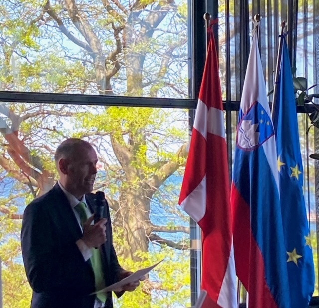 Veleposlanik stoji z mikrofonom v rokah in nagovarja povabljence, desno zadaj so danska, slovenska in evropska zastava