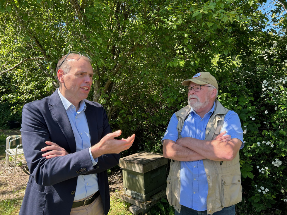 the ambassador and the beekeeper are standing in nature and talking, in the background there are trees