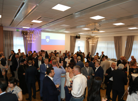 Guests mingle in the reception hall during the celebration of Slovenia’s Statehood Day. The Slovenian flag is displayed on a projection screen in the background, while attendees chat over refreshments.