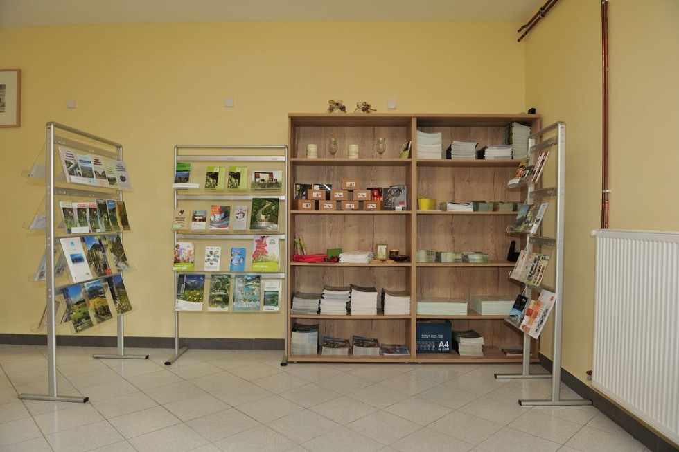 V prostoru s svetlo rumenimi stenami stoji več stojal in polic, polnih različnih brošur, knjig in zloženih papirjev. Prostor je svetel in urejen, namenjen za razstavo ali razdeljevanje informacijskih materialov.