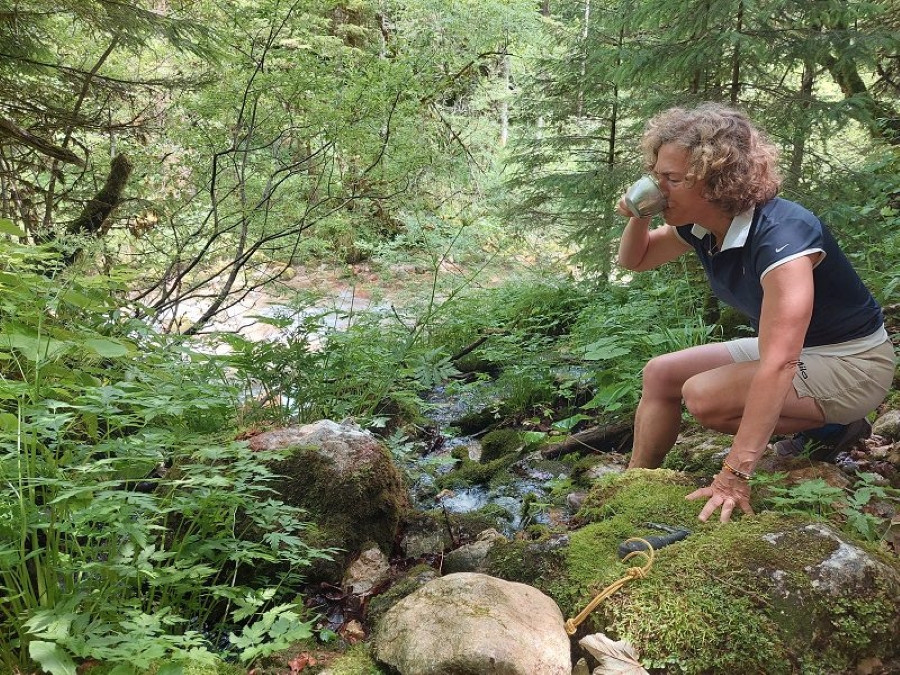 Ženska s kratkimi kodrastimi lasmi sedi na skali ob gozdnem potoku, obkrožena z zelenjem. Drži skodelico in pije. Okoli so kamni, mah in listje, v ozadju pa gosto listje in drevesa.