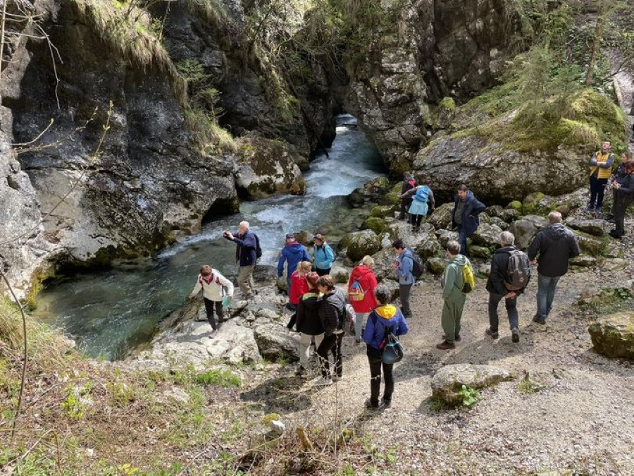 Skupina ljudi se zadržuje ob kamniti poti ob bistri gorski reki, ki teče med skalnatimi stenami in obdana z drevesi. Nekateri si ogledujejo reko, medtem ko se drugi pogovarjajo ali fotografirajo naravno okolico.