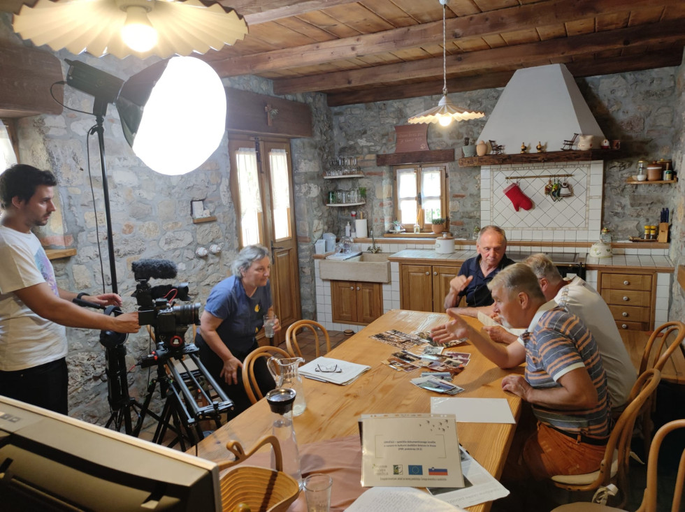 V sobi za leseno mizo sedi več oseb, ki se med seboj pogovarja. Pred njimi so postavljene luči in kamere, ki pogovor snemajo. 