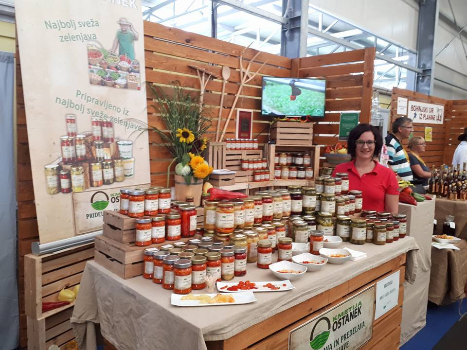 Na tej sliki je prikazan sejem ali tržnica, kjer stojita prodajna prostora z izdelki. V ospredju je stojalo, kjer Damjana Ostanek prodaja različne domače izdelke v kozarcih, kot so omake, marmelade ali vložene zelenjave.