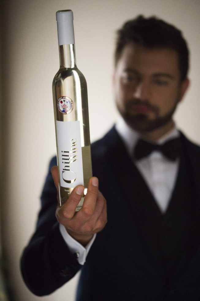 Moški v temni obleki drži steklenico belega vina z napisom »Chilli Wine«. Ozadje je zamegljeno, poudarek je na steklenici, ki ima nagradno nalepko s tekmovanja.