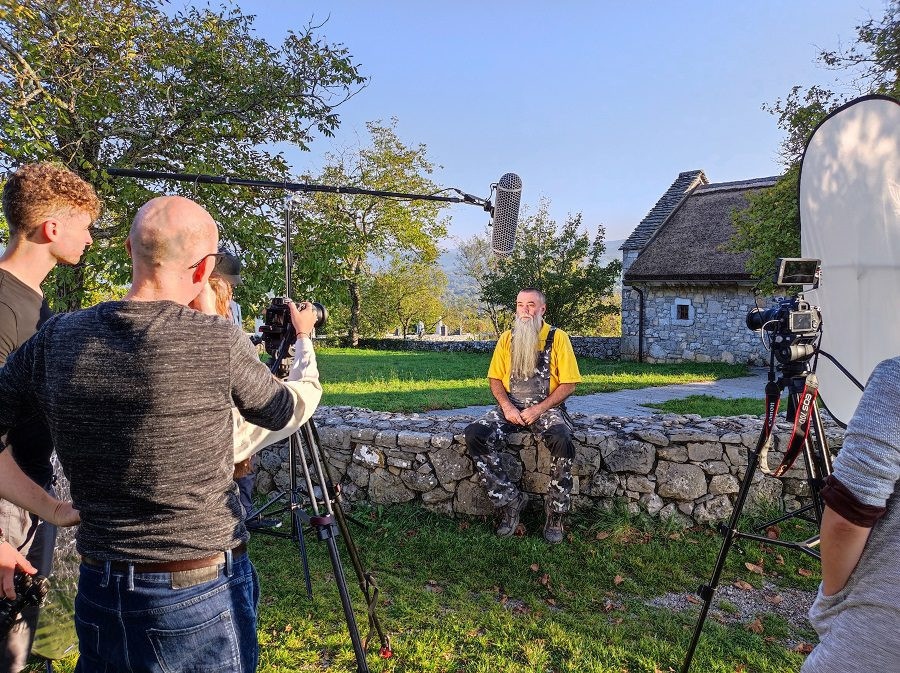 Pogled na osebo, ki govori v kamero. Nad njegovo glavo je mikrofon. V ozadju je narava. 
