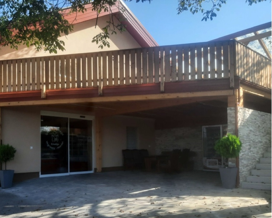 Hiša kmetije Vogrinčič-Hanžel. Ta slika prikazuje vhod v objekt s teraso nad njim. Streha terase je izdelana iz lesa z lesenimi ograjami. Pod teraso je vhod v zgradbo, verjetno glavna vrata s steklenimi drsnimi vrati.