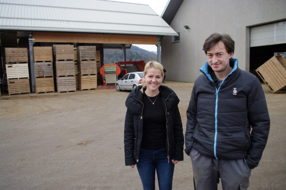 Dve osebi stojita na dvorišču pred poslopjem in se fotografirata. 