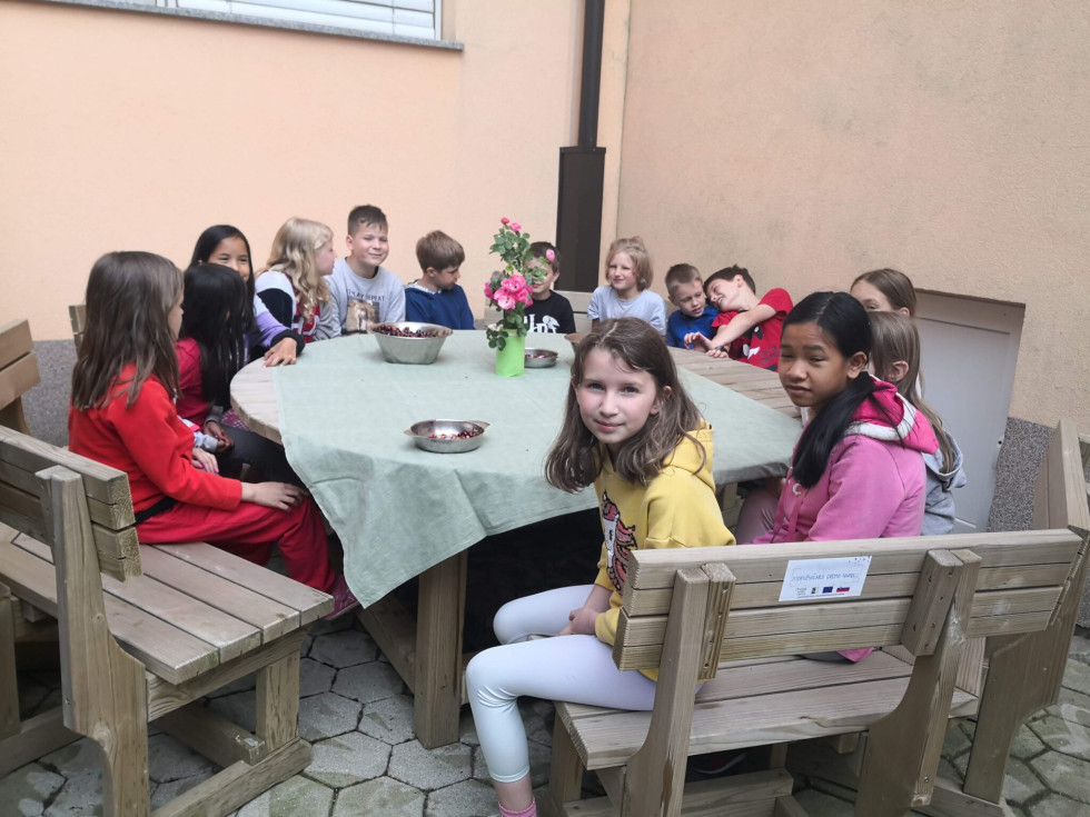 Skupina otrok sedi okoli velike lesene mize na dvorišču ob stavbi. Miza je pokrita z zelenim prtom, na njej so rože in nekaj skledic. Otroci se družijo in pogovarjajo v prijetnem zunanjem okolju.