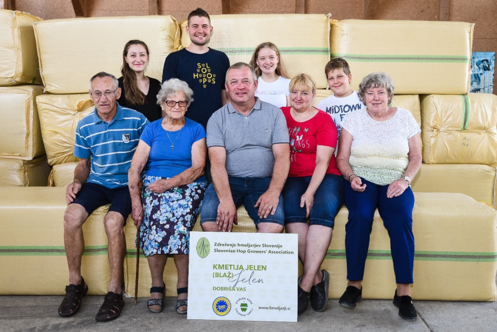 Skupinska fotografija večgeneracijske družine na kmetiji Jelen. Osem oseb sedi in stoji pred zloženimi balami sena v hlevu. Spredaj držijo tablo z napisom »Kmetija Jelen« in logotipi združenja hmeljarjev Slovenije.