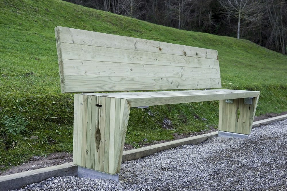 Fotografija prikazuje moderno leseno klop z naslonom, postavljeno ob urejeno pot iz drobnega kamenja. Klop je narejena iz svetlega, obdelanega lesa in je nameščena ob rahlem pobočju z zeleno travo.