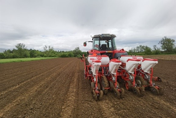 Rdeč traktor z večvrstno belo-rdečo sejalnico na oranici. Nebo je oblačno, v ozadju so drevesa.