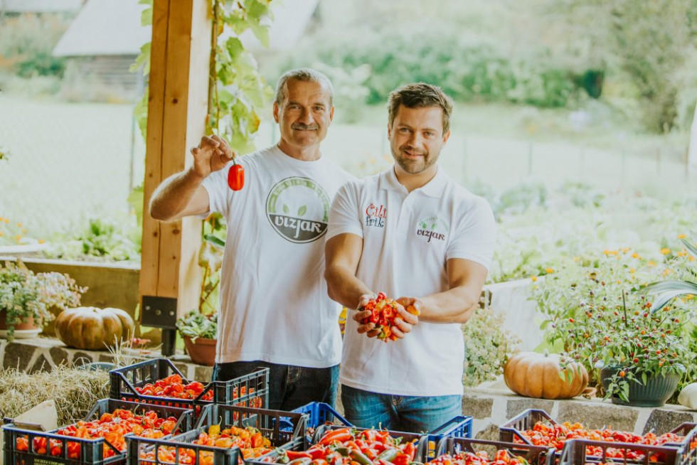 Oče in sin stojita pod nadstreškom, obkrožena z zaboji rdečih čilijev. Oba nosita majici z logotipom kmetije Vizjak.