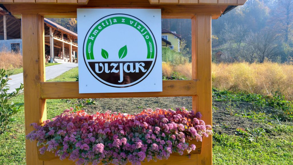 Na lesenem stojalu je bela tabla z logotipom "Vizjak – kmetija z vizijo". Pod tablo je korito s cvetočimi rožami, v ozadju pa je podeželska pokrajina.