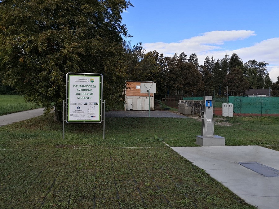 Na travnati površini ob robu ceste stoji tabla z napisom "Postajališče za avtodome motorhome stopover". Ob tabli je steber za priklop na elektriko ali vodo. V ozadju so drevesa, stavbe in ograjeni športni objekti.