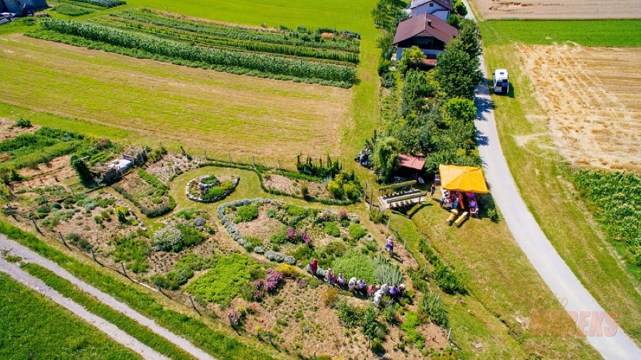 Na fotografiji je zračni posnetek podeželskega območja z urejenim zeliščnim vrtom in nekaj stavbami. Okoli vrta so polja in drevje, kar poudarja mirno podeželsko okolje.V desnem delu slike se vidijo tudi gospodarska poslopja in nekaj parkiranih vozil.