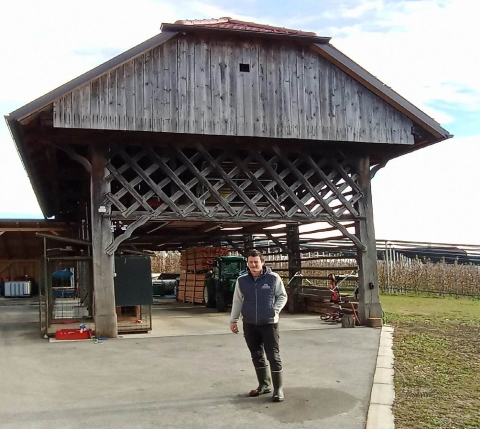 Pred veliko leseno zgradbo s tradicionalno streho stoji Bojan Frešer. Zgradba ima odprt prostor pod streho, v katerem so shranjeni leseni zaboji in traktor. Okoli je asfaltirana površina in polje.
