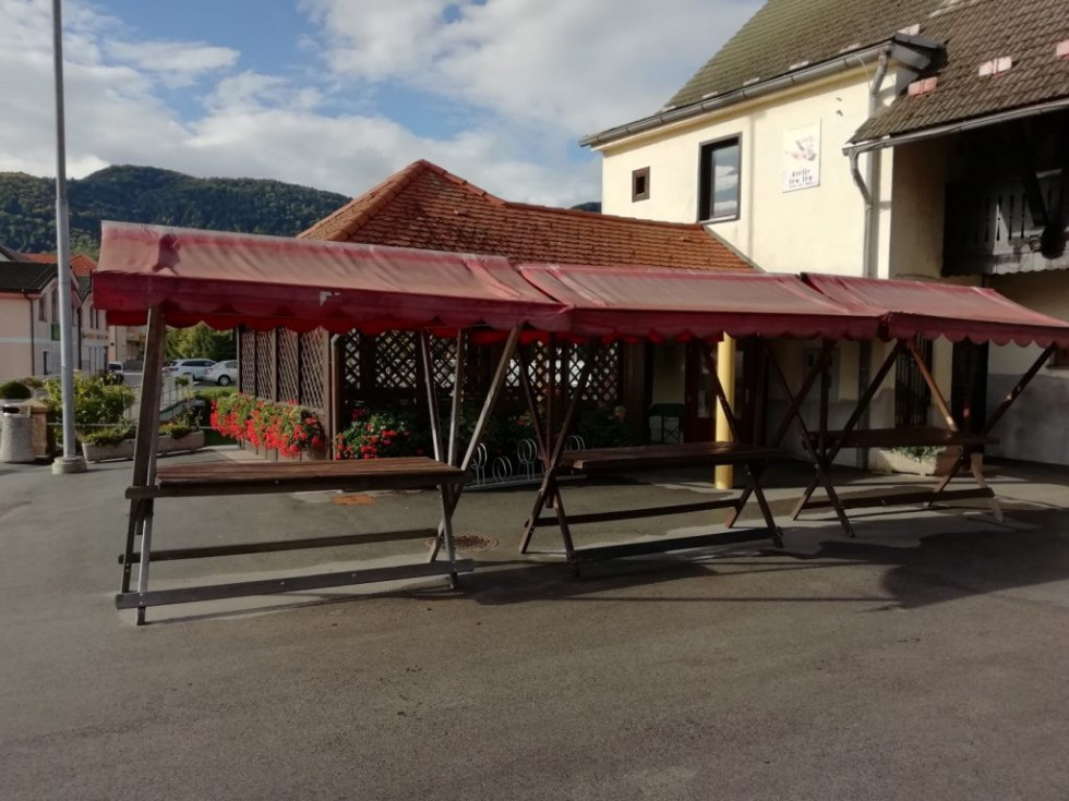 Na asfaltirani površini pred staro stavbo z rumenimi stenami stojijo prazne tržne stojnice z rdečimi platnenimi nadstreški.
