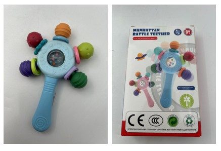 Otroška ropotuljica z grizalom "Manhattan Rattle Teether", svetlo modre barve, s petimi barvnimi grizalnimi kroglicami okoli osrednjega dela s prozornim okencem. Izdelek je pakiran v kartonski embalaži z napisom »MANHATTAN RATTLE TEETHER«.