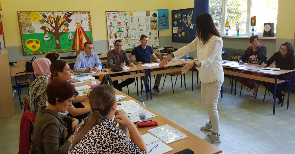 Ženska stoji na sredini razreda in razlaga odraslim, ki sedio v šolskih klopeh.