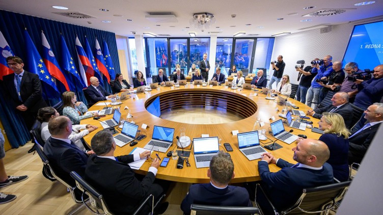 People sitting at a round table with computers in front of them in a room with slovenian and EU flags.