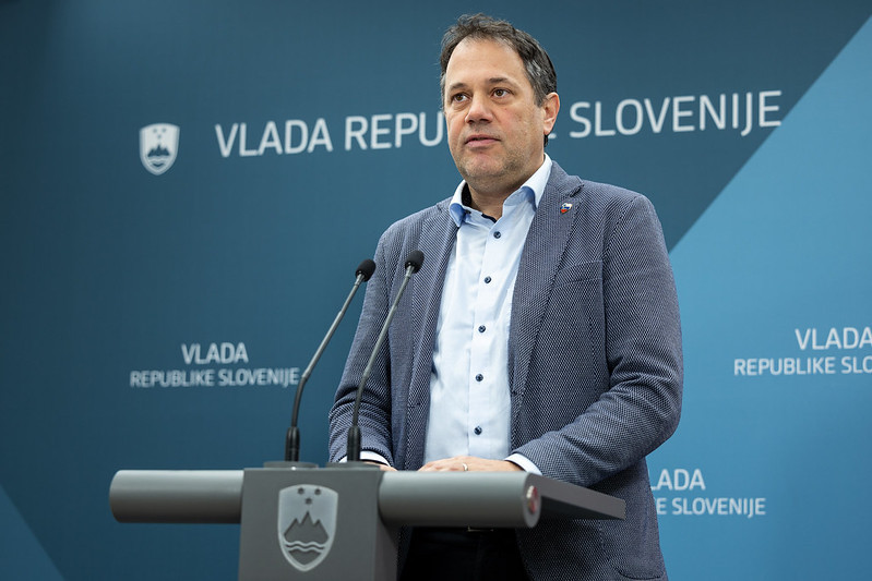 A man speaking at a press conference with "VLADA REPUBLIKE SLOVENIJE" in the background, indicating a Slovenian government event.
