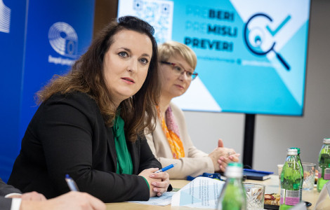 54394910930 1d096b7561 c (A professional meeting with two women sitting at a table, taking notes. A screen in the background displays a message in Slovenian: "Preberi, Premisli, Preveri." Bottled water and documents are on the table.)