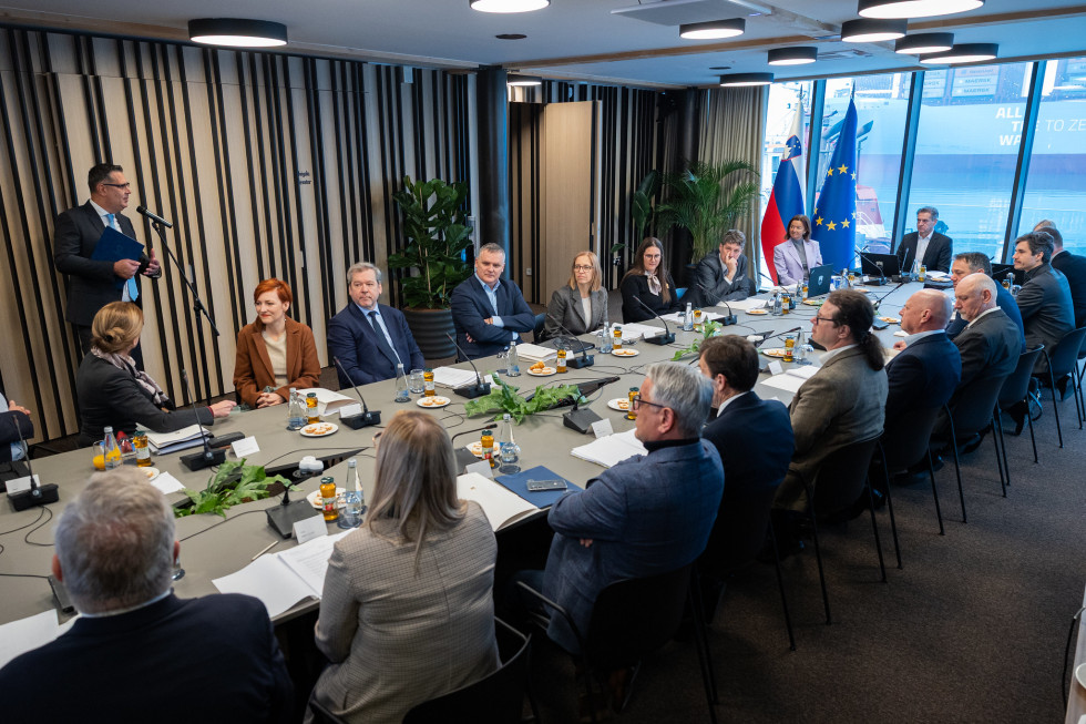 Moški in ženske sedijo za dolgo mizo. V kotu sobe slovenska in evropska zastava.