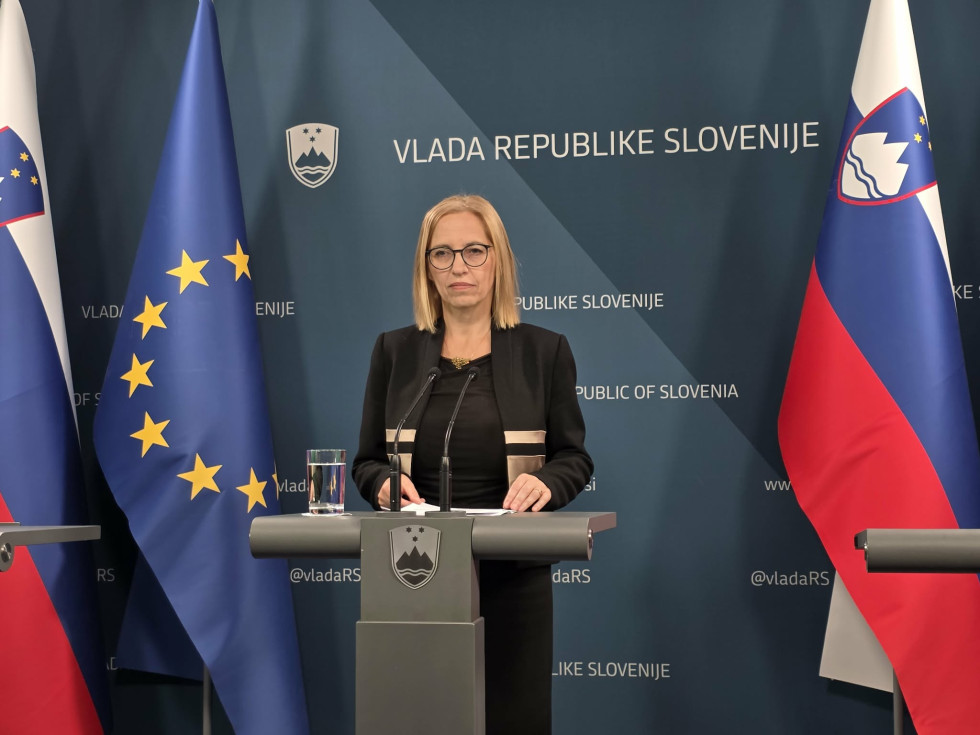 A woman stands behind a lectern, with the European and Slovenian flags behind her.
