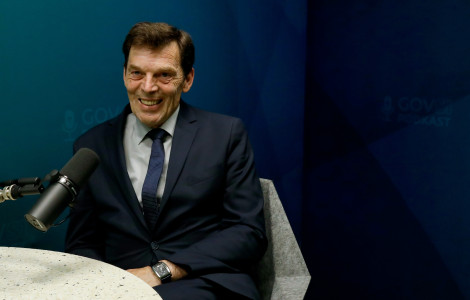AZC0500 (Moški srednjih let do starejši, v temnem poslovnem oblečnem suknjiču, srajci in kravati, sedi nasmejan pred mikrofonom v studiu z modro ozadjem. Ozadje vsebuje logotip »GOVSI PODKAST«. Moški ima kratke temne lase z nekaj sivine in nosi uro na levi roki. P)